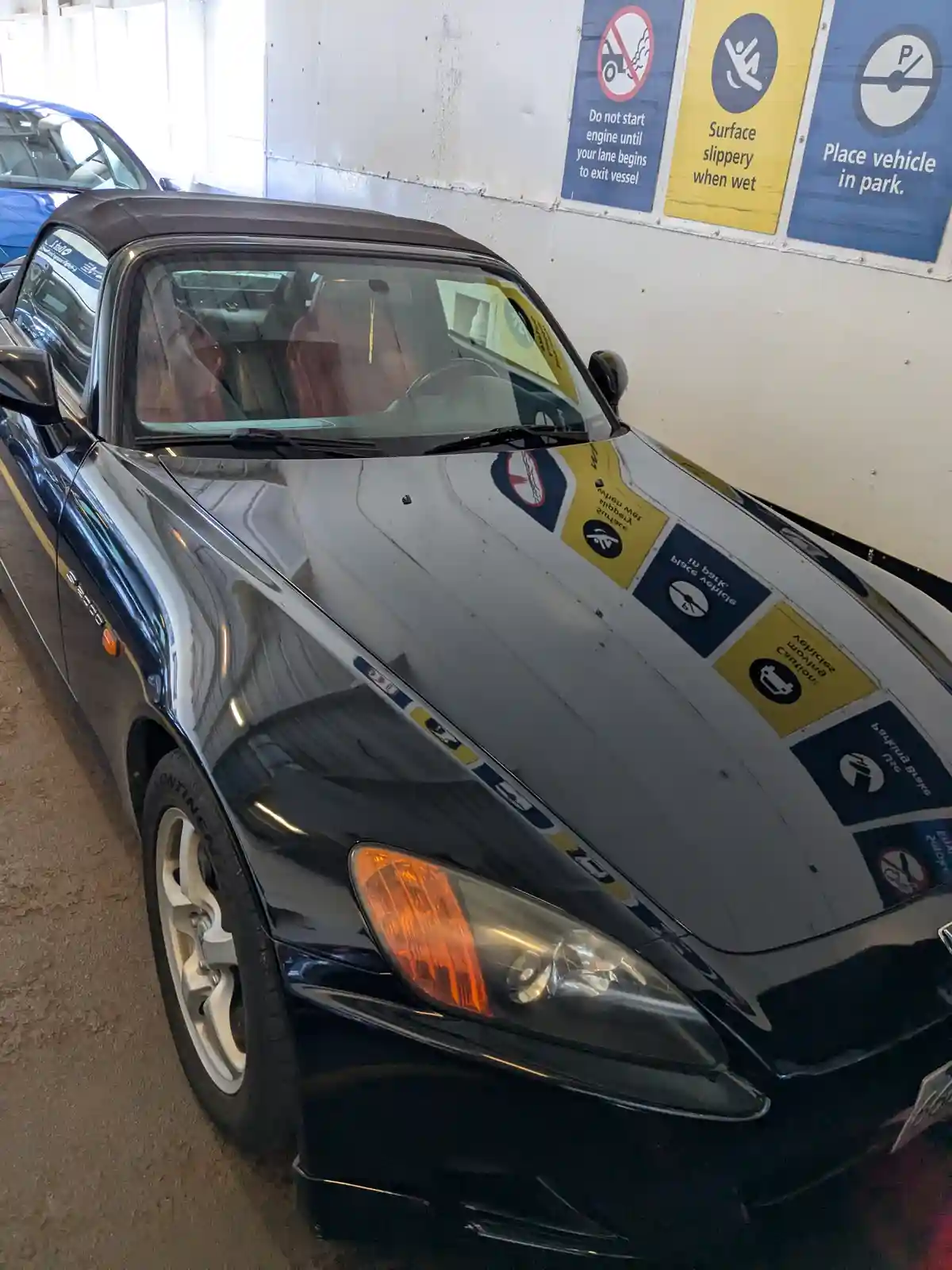 A black Honda S2000 convertible is parked indoors with safety signs from a boat ramp reflected on its hood. The signs…