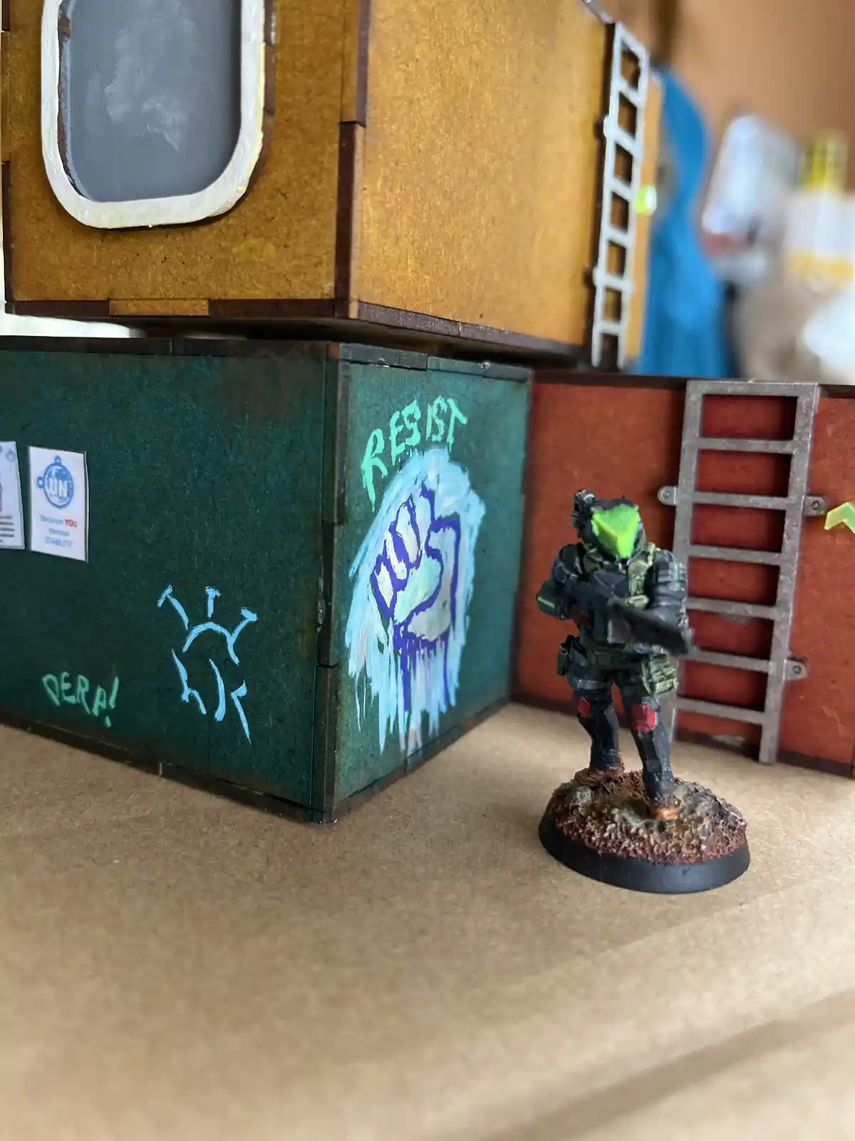 A painted miniature figure in green and black armor stands on a rocky base next to a green box with graffiti. The box has a…