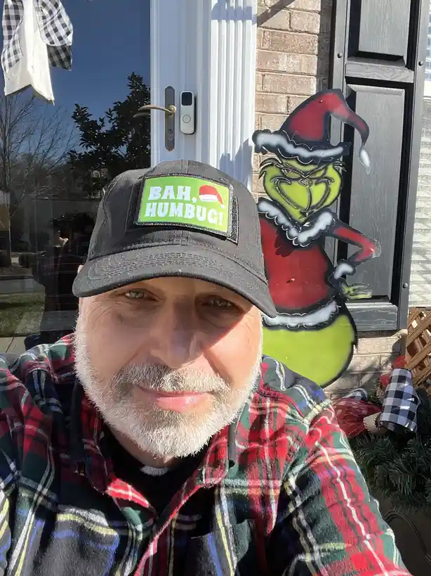 A man with a white beard and mustache wearing a black baseball cap with a green patch that reads 'BAH, HUMBUG!' and a red…