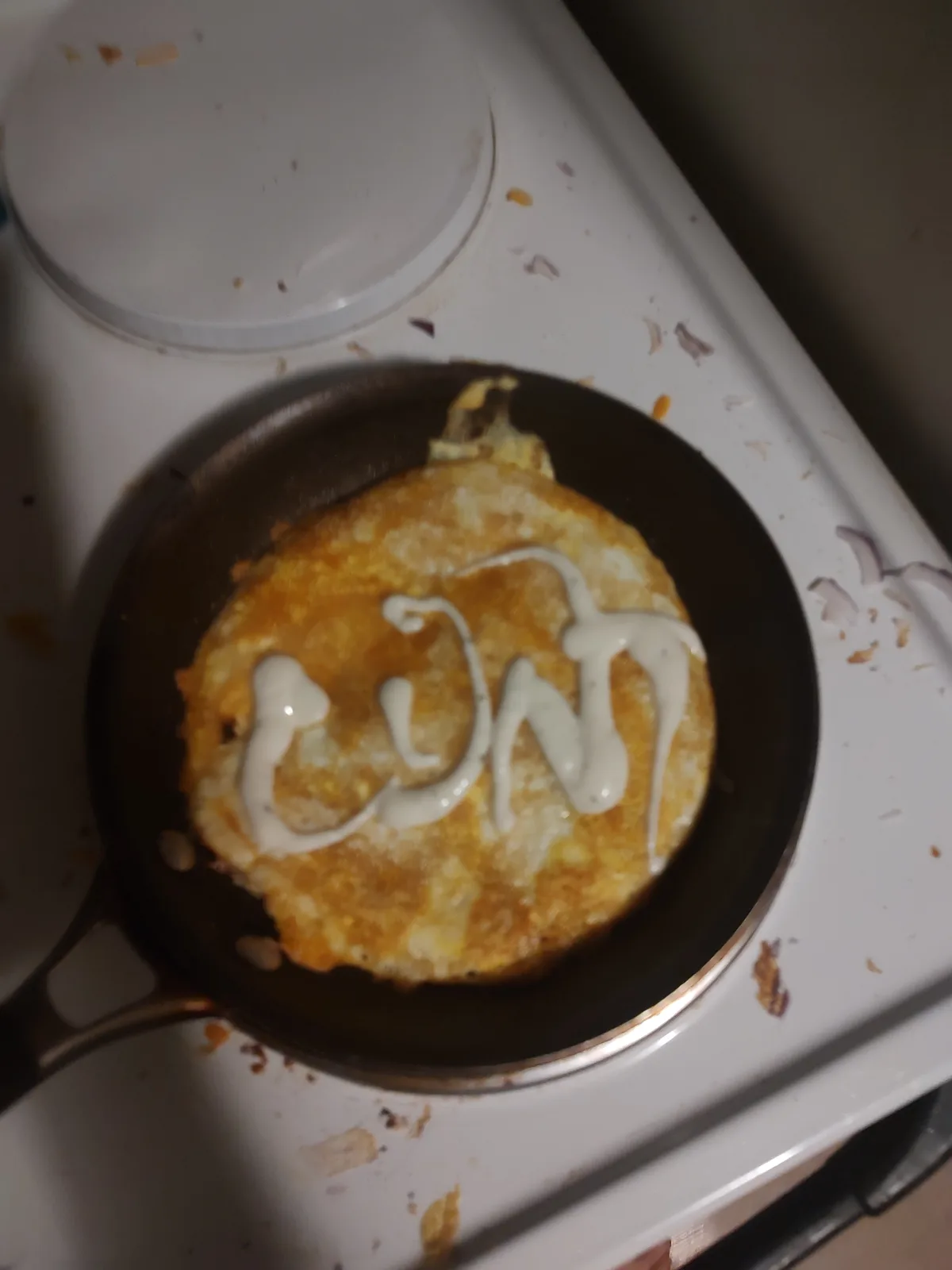 A close-up photo shows a golden-brown fried egg in a black frying pan on a white stovetop. A white sauce, likely ranch or…