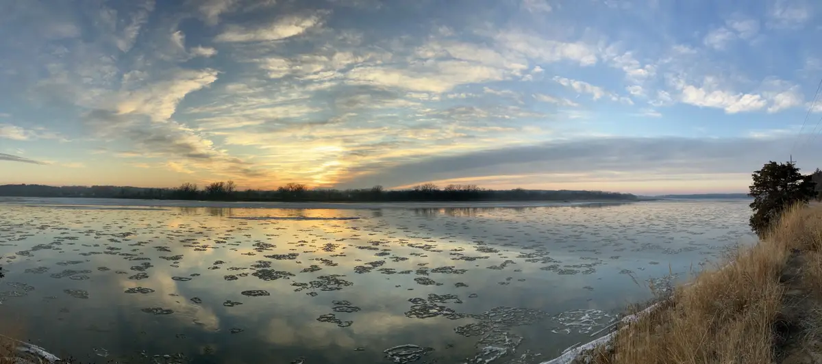 A wide-angle photo of a partially frozen river at sunrise or sunset, with ice floes scattered across the water reflecting…