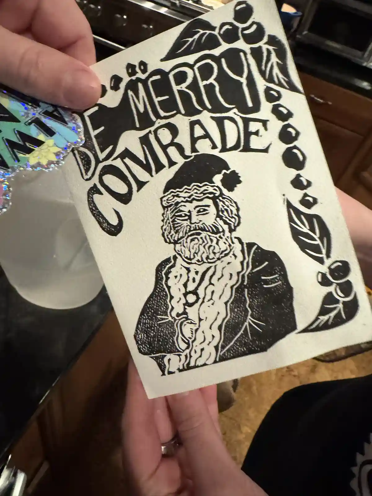 A hand holds a handmade black-and-white holiday card featuring a stylized, bearded Santa Claus wearing a hat, framed by…
