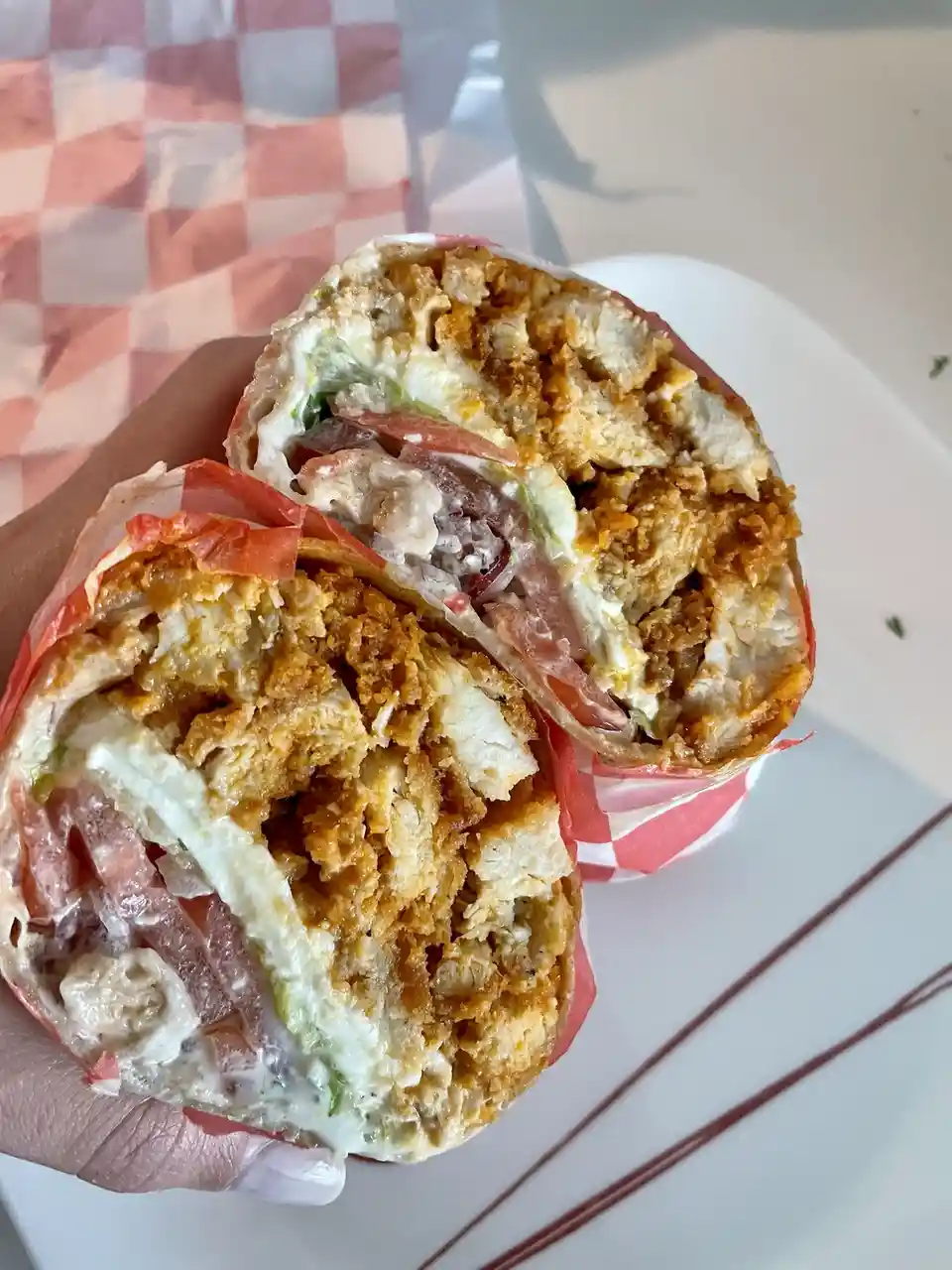 A close-up photo shows a hand holding a sandwich cut in half, revealing crispy fried chicken, tomato slices, lettuce, and…