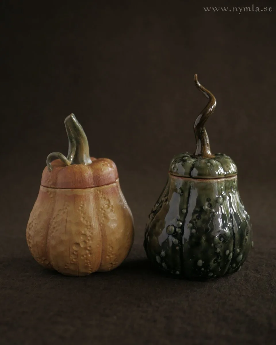 Two ceramic containers shaped like pumpkins with lids, one tan and one green, sit side by side on a dark surface. The tan…