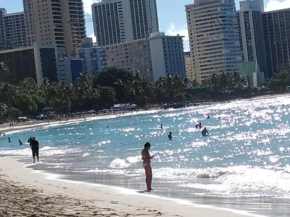 A woman in a bikini stands at the water's edge on a sunny beach with tall city buildings in the background. Other beachgoers…
