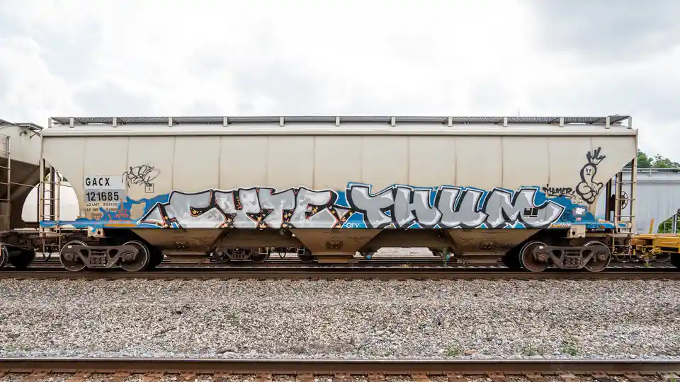 A side view of a white and brown railroad hopper car covered in graffiti, prominently featuring the word 'EXTREMELY' in…