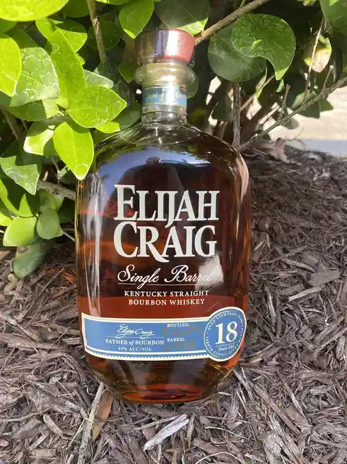 A bottle of Elijah Craig Single Barrel Kentucky Straight Bourbon Whiskey sits upright in brown wood chip mulch, partially…
