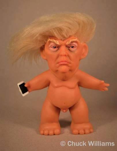 A small, nude, orange plastic figurine resembling a troll with a large blonde wig and a scowling face, holding a small black…