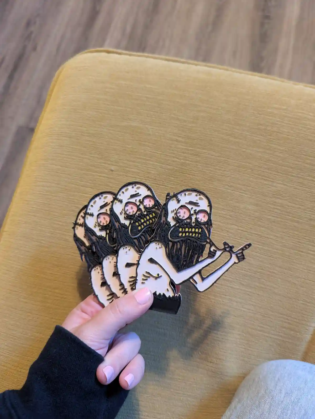 A hand holds a stack of laser-cut, cartoonish skull-shaped pins over a yellow cushion. The skulls have pink eyes, black…