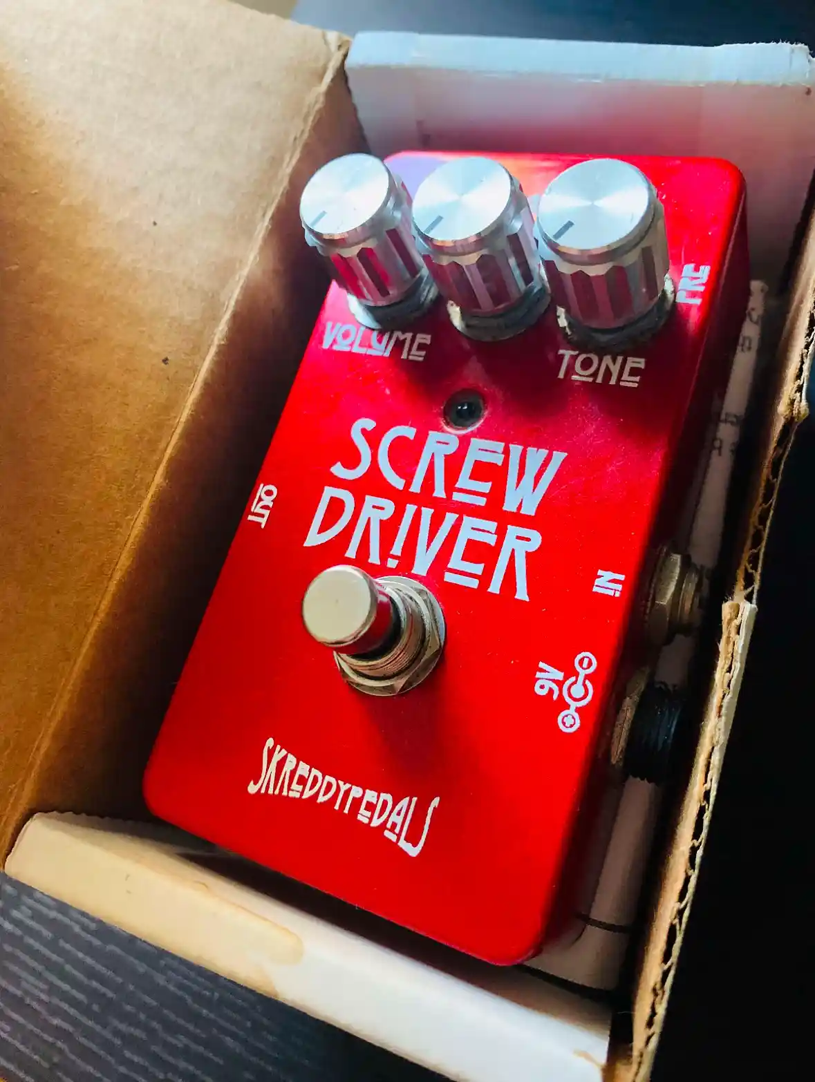 A red guitar effects pedal labeled 'SCREW DRIVER' by 'SKREDDYPEDALS' sits in a cardboard box. It has three silver knobs…