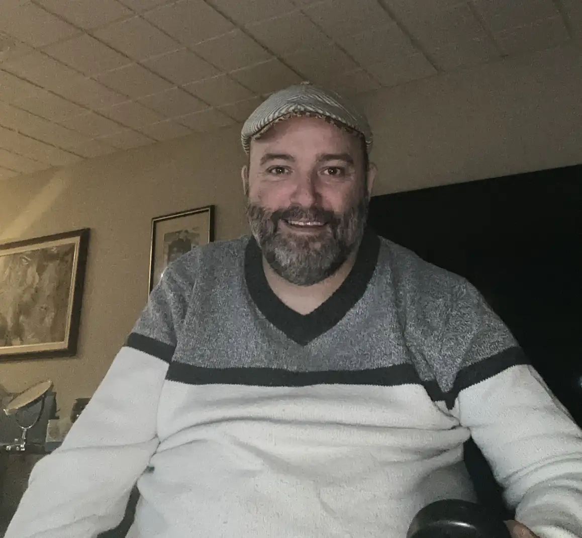 A man with a beard and a grey flat cap smiles at the camera while wearing a grey and white striped sweater. Behind him, a…