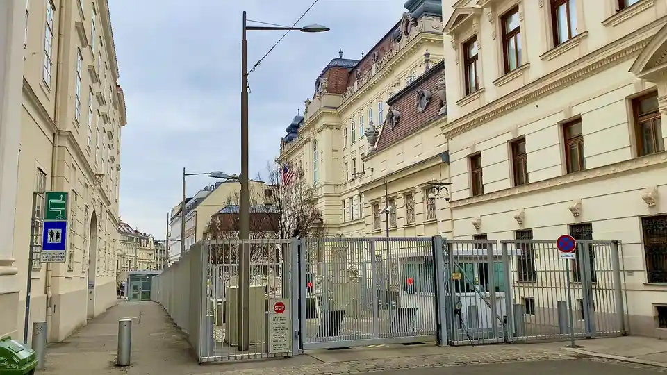 An exterior view of a large, ornate, light-colored building with a high fence and gate in front. An American flag is visible…