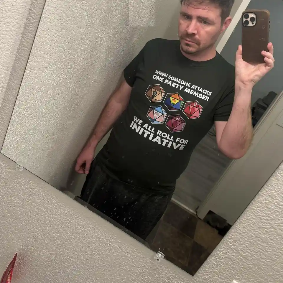 A man takes a mirror selfie holding a smartphone, wearing a black t-shirt with white text and colorful dice graphics. The…