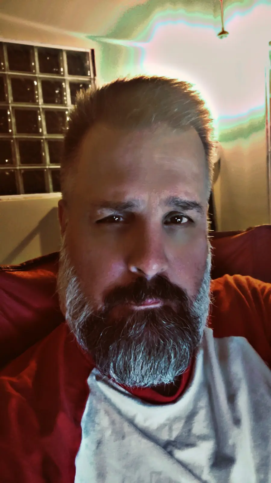 A man with a graying beard and short hair sits in a red chair, wearing a red and white shirt, looking directly at the camera…
