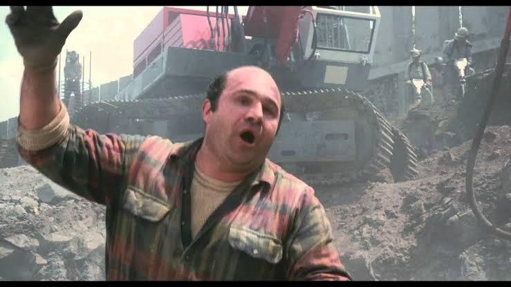 A man in a plaid shirt with a shocked expression raises his hand, standing in front of construction equipment and workers…