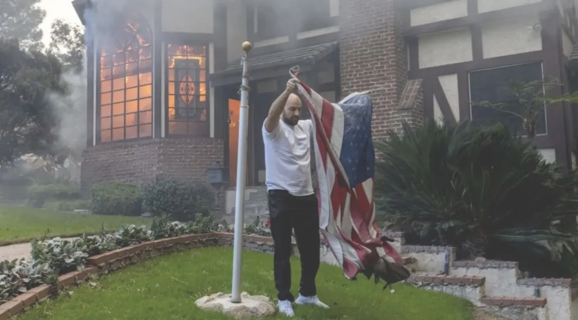A man stands in a grassy yard in front of a house engulfed in flames and smoke, holding an American flag and preparing to…