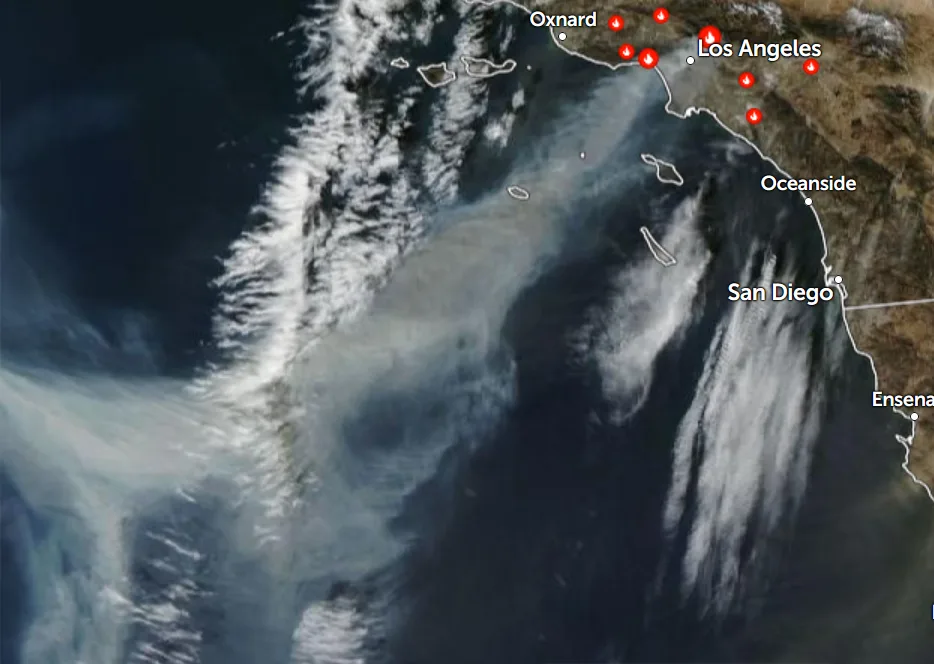 A satellite image shows a large plume of smoke drifting over the coast of Southern California, with red fire icons overlaid…
