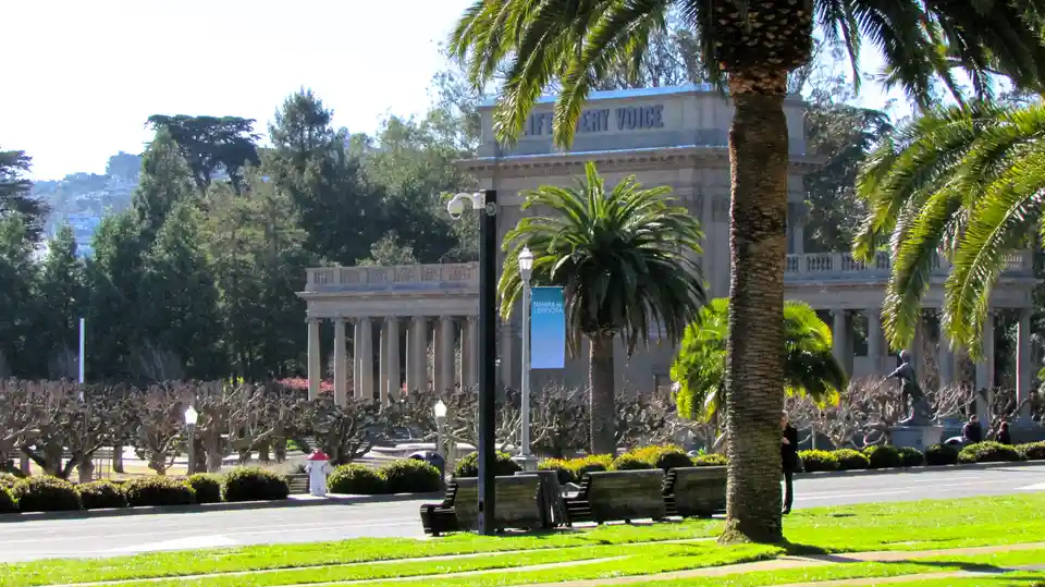 A sunny park scene with palm trees, benches, and a classical building in the background. The building's facade has the words…