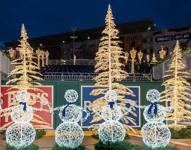 A nighttime photo shows four illuminated snowman figures made of white string lights, standing in front of large, lit-up…