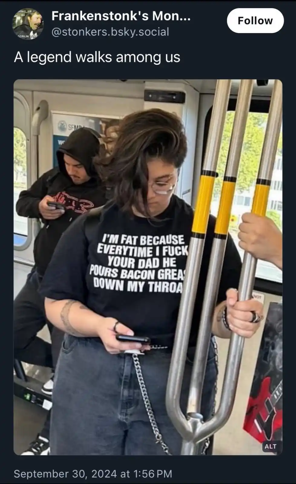 A person on public transit wearing a black t-shirt with the text 'I'M FAT BECAUSE EVERYTIME I FUCK YOUR DAD HE POURS BACON…