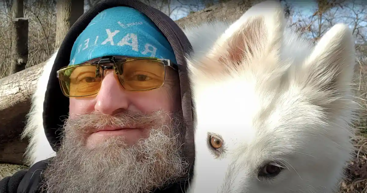 A man with a long beard, wearing a blue headband and yellow-tinted glasses, takes a selfie outdoors with a white dog. The…