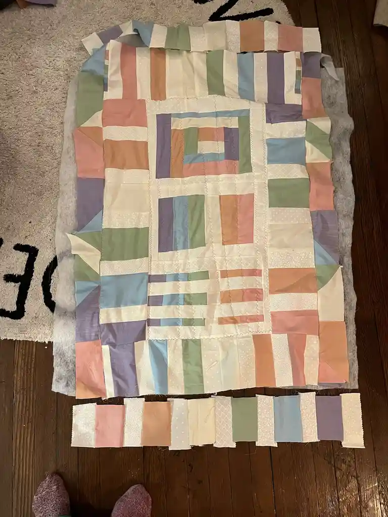 An overhead photo shows a partially assembled patchwork quilt laid out on a wooden floor next to a rug, with mismatched…