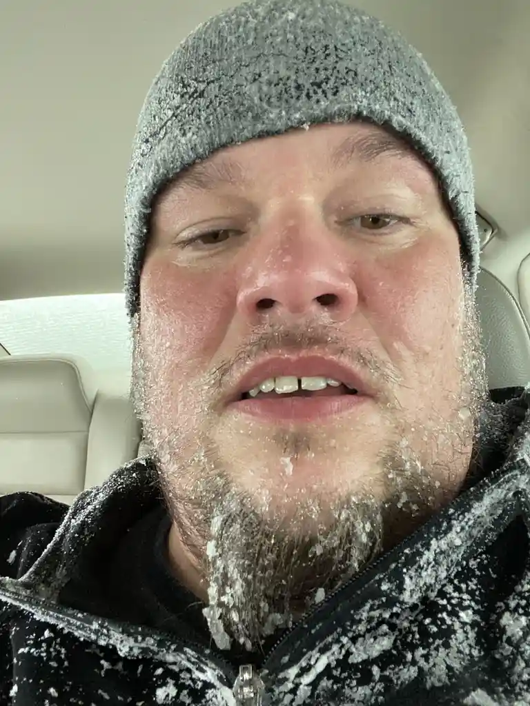 A man inside a car has frost and ice crystals covering his face, beard, hat, and jacket, indicating extreme cold. He looks…