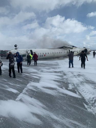 A damaged airplane lies on a snowy tarmac with smoke rising from its fuselage, while several people in winter clothing stand…