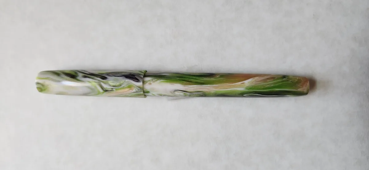 A horizontally placed pen with a swirling, marbled pattern of green, white, black, and orange sits on a plain white textured…