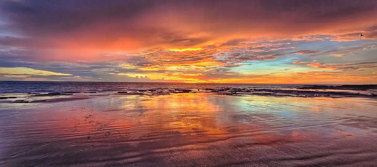 A panoramic photo of a stunning sunset over a wet beach, with vibrant orange, pink, and purple clouds reflected in the…