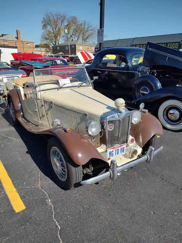 A vintage beige and brown MG TD roadster is parked at a car show, featuring a skull ornament on its hood and an Ohio license…