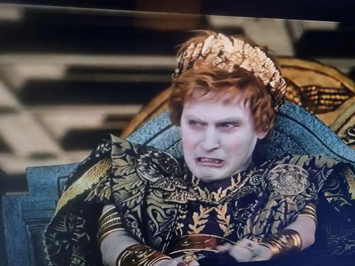 A man with red hair wearing an ornate golden crown and elaborate black-and-gold royal robes sits on a throne, displaying an…