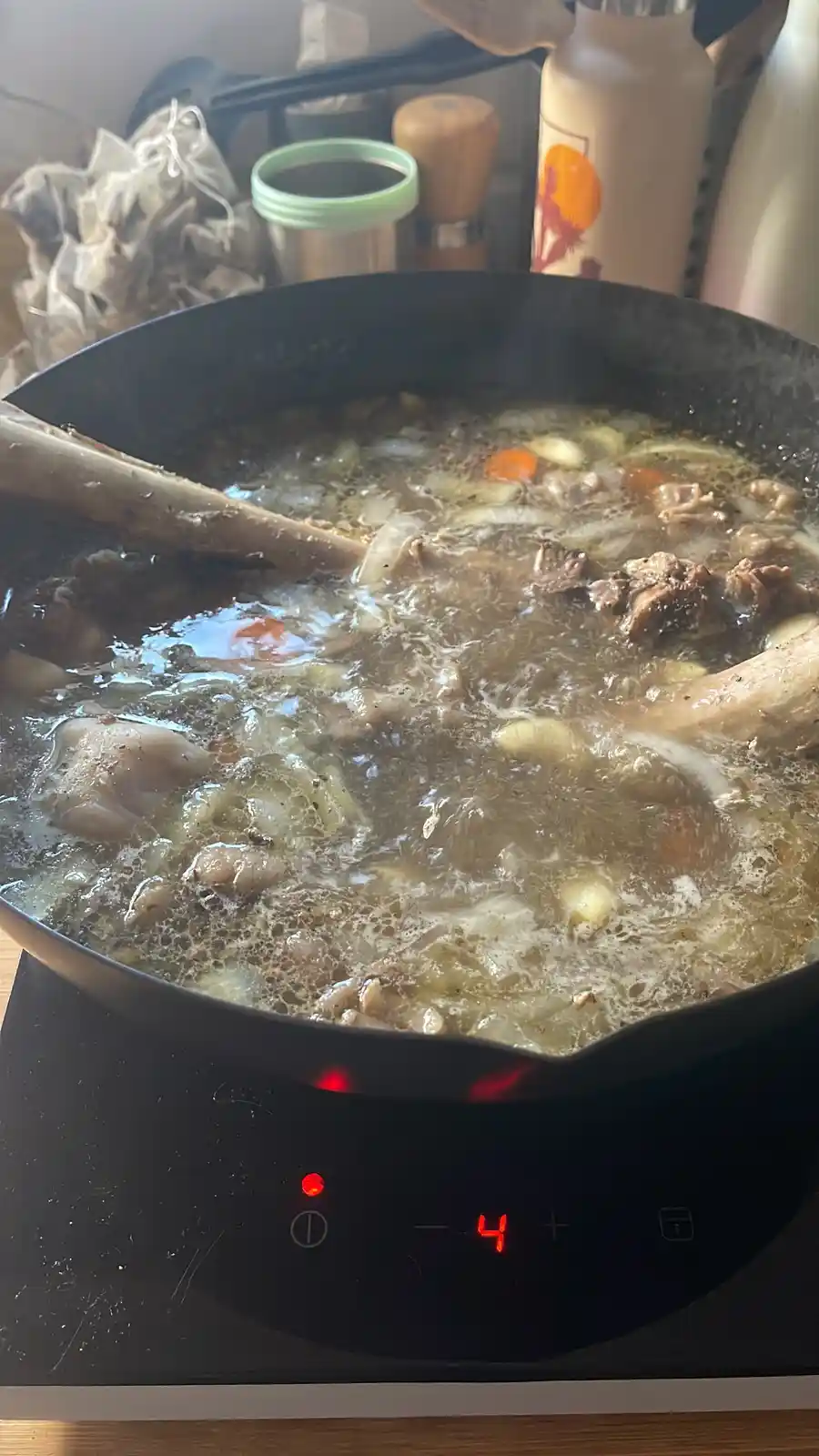A close-up photo shows a black frying pan on an electric stove set to level 4, boiling a dark, bubbling stew with large…