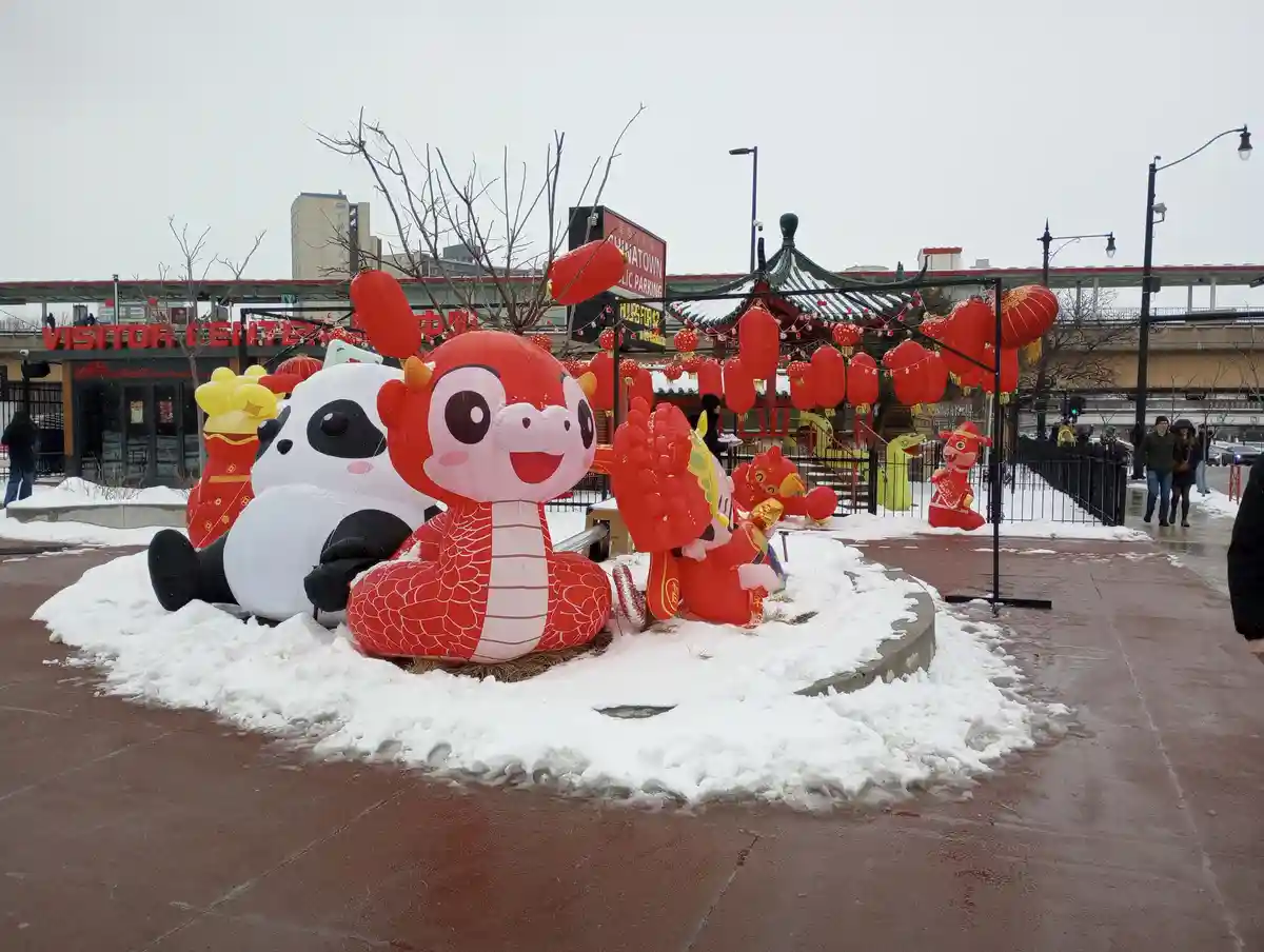 A snowy outdoor scene features large inflatable Chinese New Year decorations, including a red dragon and a panda, surrounded…
