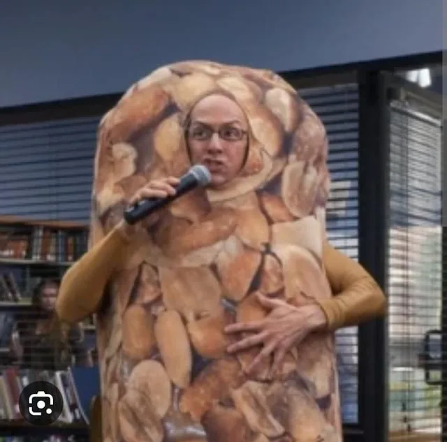 A person wearing a full-body costume resembling a large, textured bag of roasted nuts stands indoors holding a microphone…