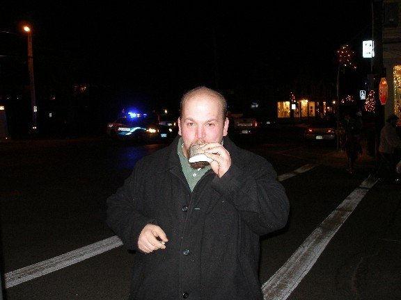A bald man in a dark coat stands on a street at night, holding a cup to his mouth as if drinking, with police lights visible…