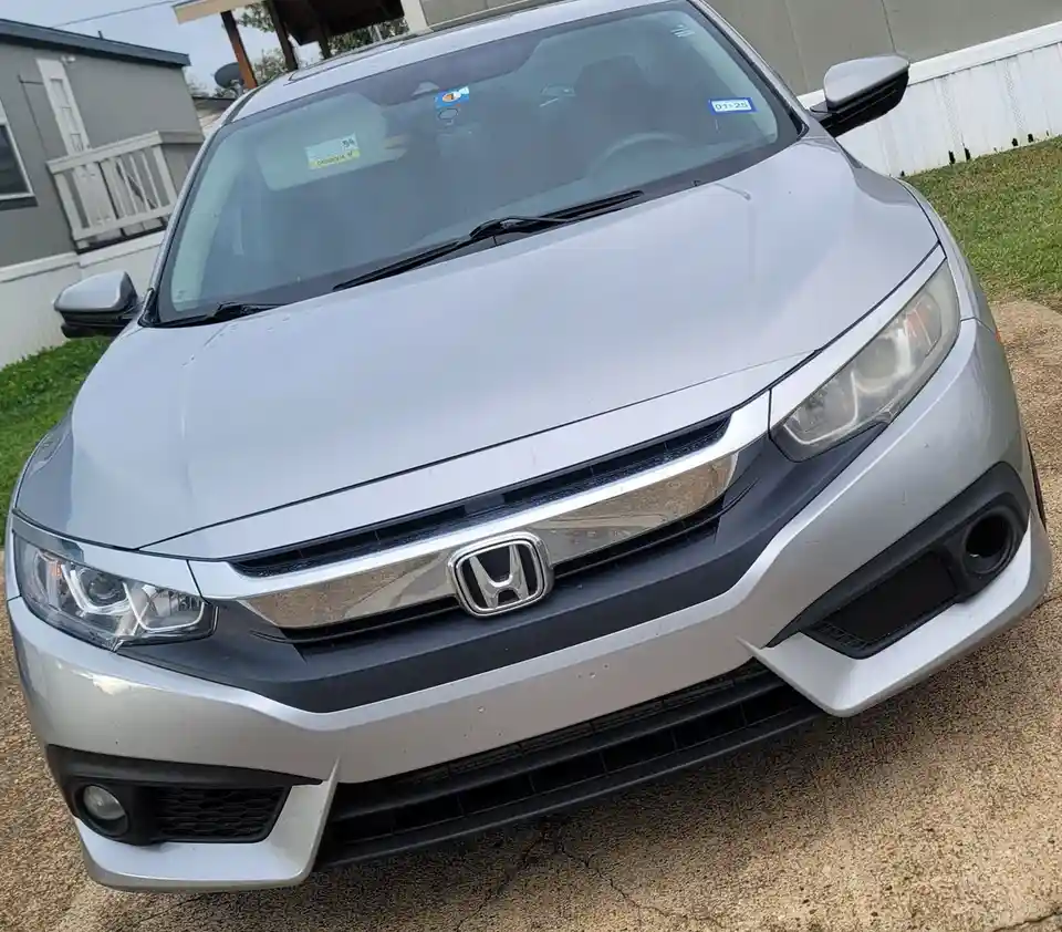 A silver Honda Civic is shown from a front-facing angle, parked on a gravel driveway with a house and fence in the…