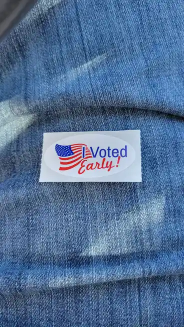 A close-up photo shows a white oval sticker with an American flag design and the text 'I Voted Early!' stuck on blue denim…