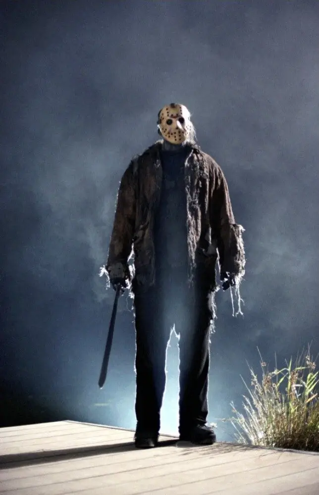 A full-body shot of Jason Voorhees from the 'Friday the 13th' franchise, standing on a wooden dock at night, wearing his…
