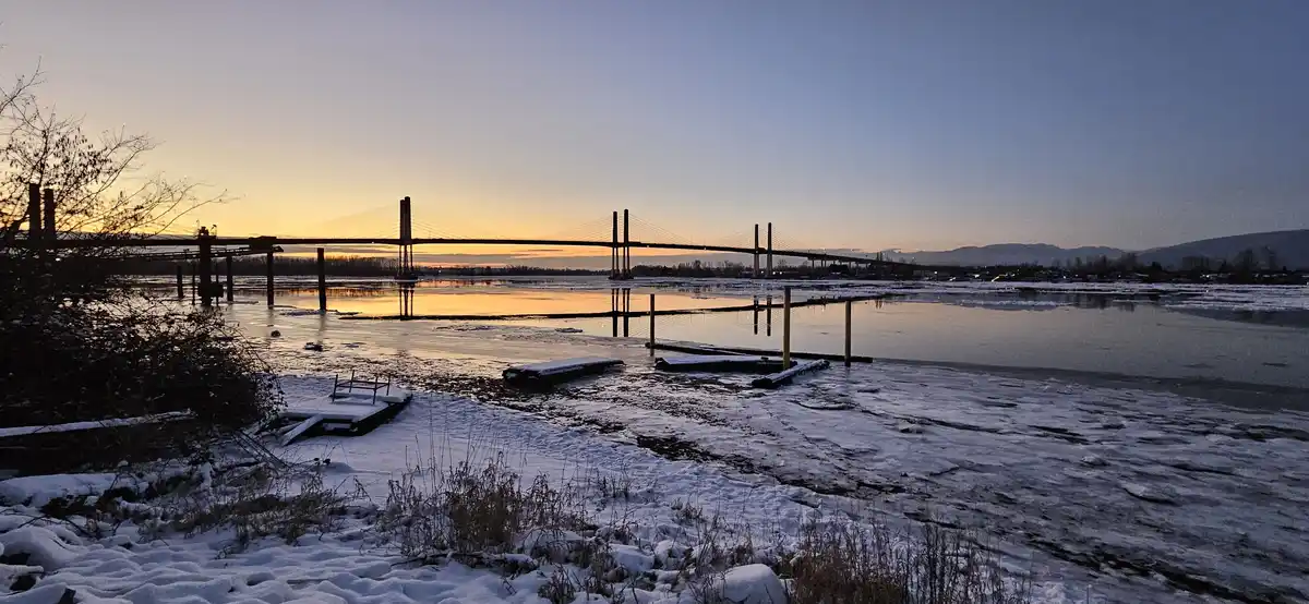 A serene winter scene at dusk shows a large cable-stayed bridge spanning a partially frozen river, with snow covering the…