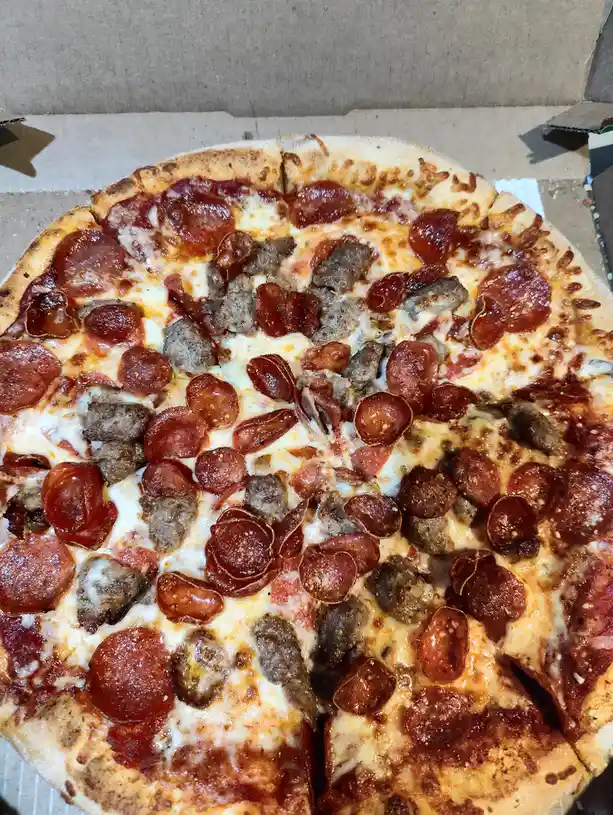 A close-up photo of a pizza in a cardboard box, visibly split into two halves: one half covered with pepperoni slices and…