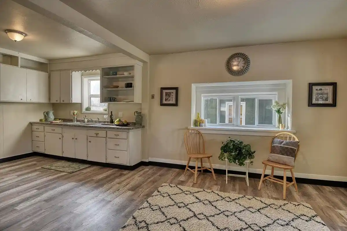 A staged photo of a beige-walled room with white cabinets, a sink, and a window, but no stove, oven, or refrigerator…