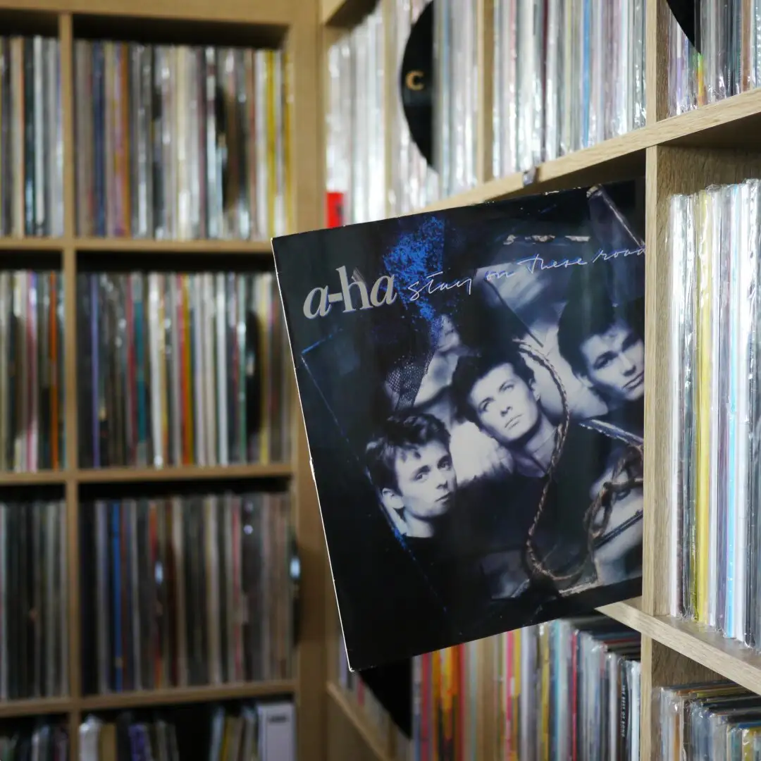 A close-up photo shows the album cover of 'a-ha' titled 'Stay on These Roads' leaning out from a wooden shelf filled with…