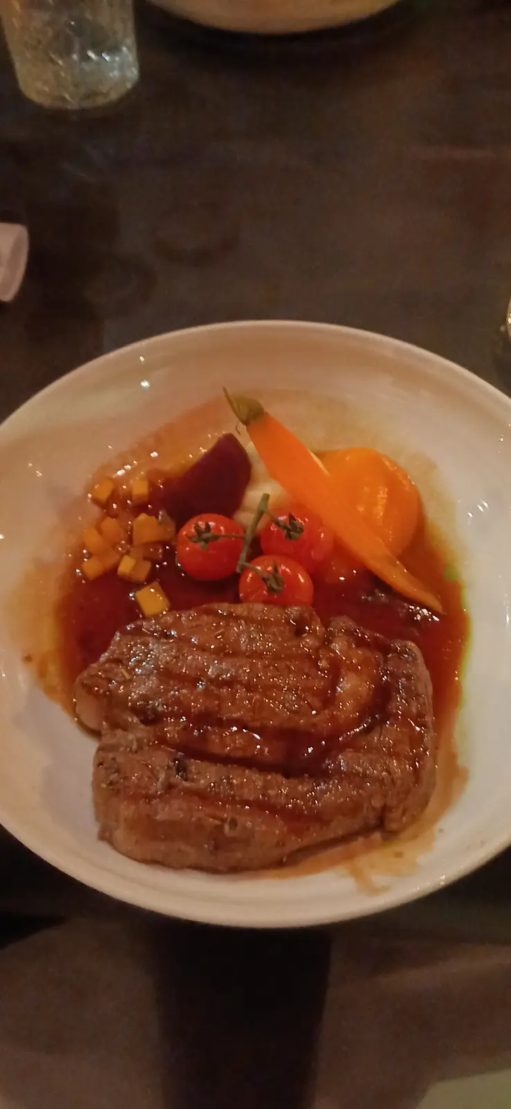 A close-up photo of a plated meal featuring a thick, seared meat patty covered in a red sauce, surrounded by cherry…