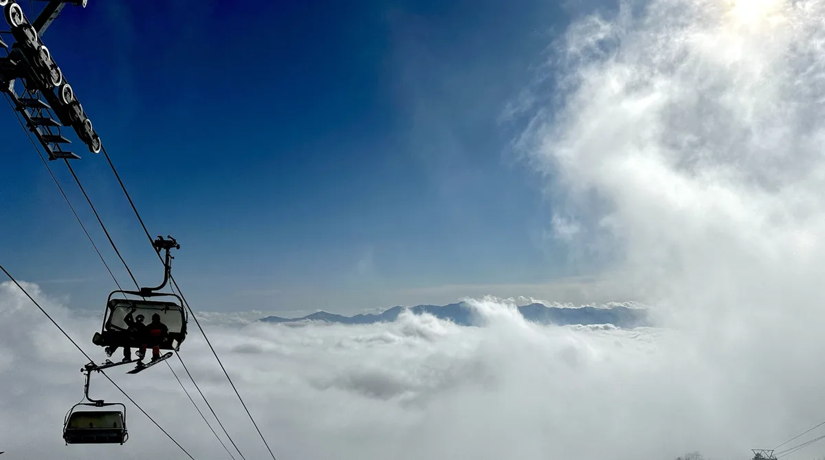 A ski lift chair with two people, one holding a snowboard, is suspended high above a thick layer of clouds, with distant…