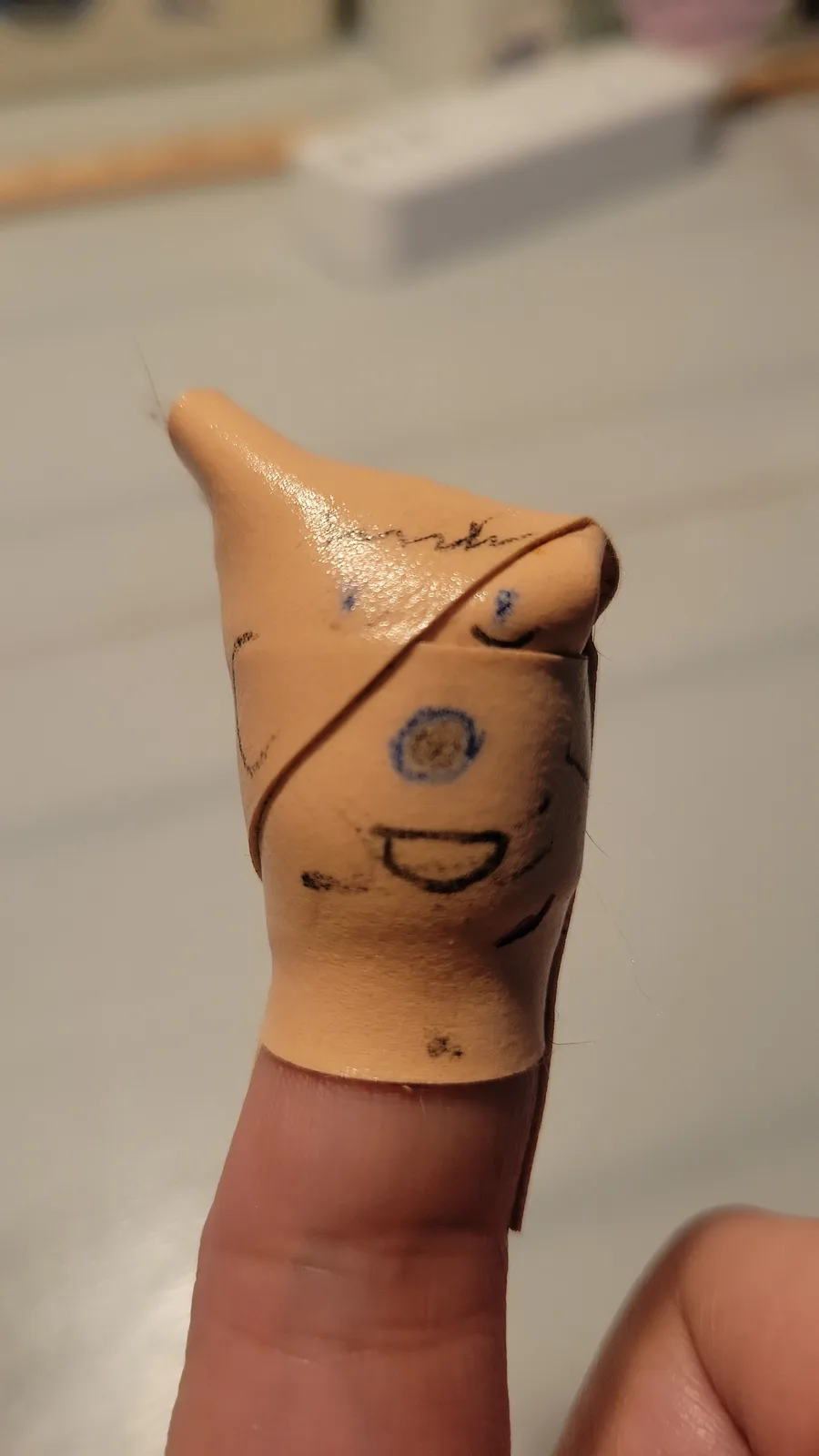 A close-up of a human finger wearing a beige, flesh-toned finger puppet made of flexible material. The puppet has a crudely…