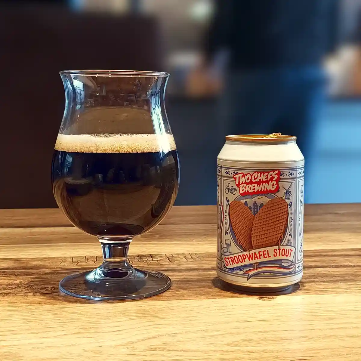 A dark stout beer in a tulip glass sits next to its can, which is labeled 'Two Chefs Brewing Stroopwafel Stout' and features…