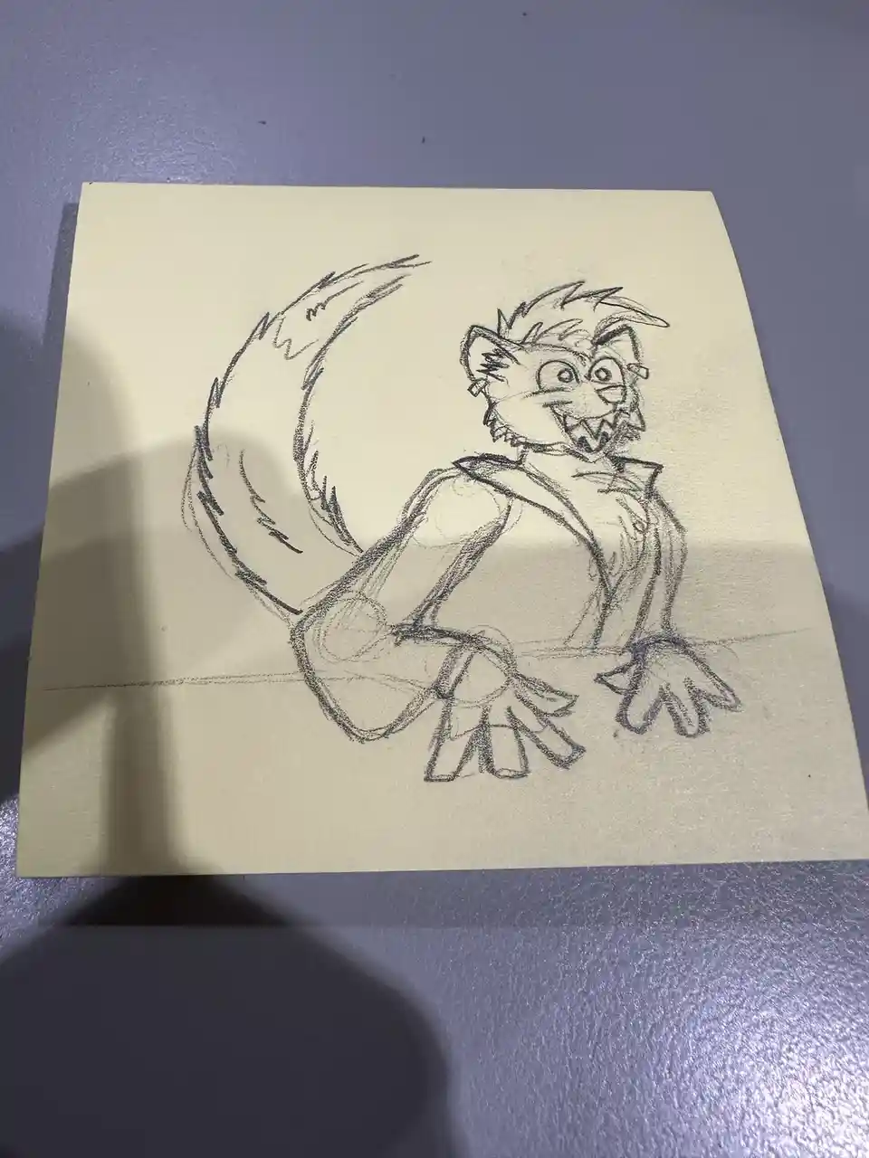 A pencil sketch on yellow paper shows a cartoonish anthropomorphic animal with spiky hair, a large bushy tail, and a wide…