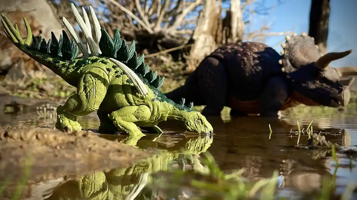 Two plastic dinosaur toys, a green stegosaurus in the foreground drinking from water and a dark triceratops in the…