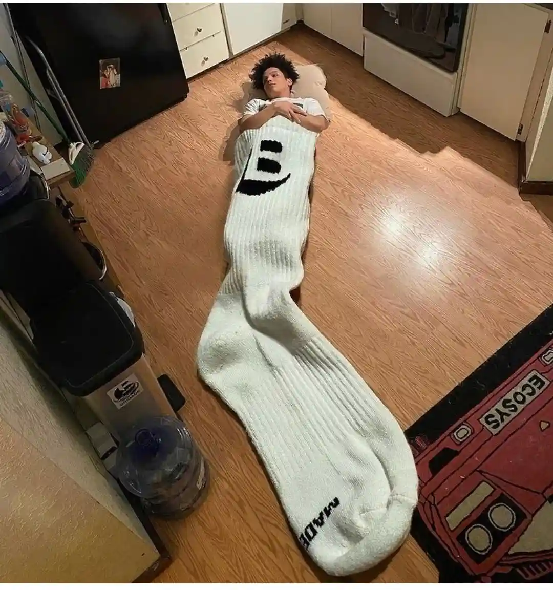A person is lying on a wooden floor in a kitchen, wrapped in an extremely large white sock that covers their entire body…